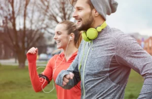 Music-and-workout-blog - RecoverRx A man and a woman are jogging together outdoors. The man is wearing a gray long-sleeve shirt, a beanie, and yellow headphones, while the woman is wearing a red jacket and earphones. Both are smiling and enjoying their run with trees and a park in the background.