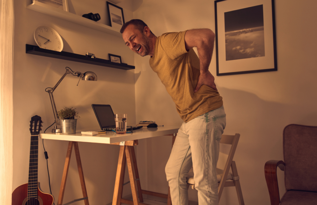 A man in a yellow t-shirt stands beside a home office desk, clutching his lower back in pain related to pelvic health, with a grimace on his face, in a warmly lit room with